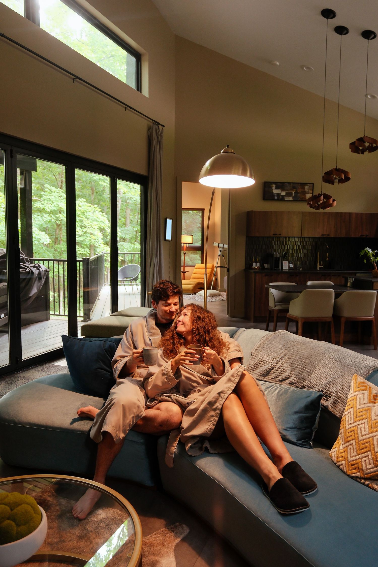 Couple in robes relaxing on a blue sofa in a modern luxury cabin living room with forest views.