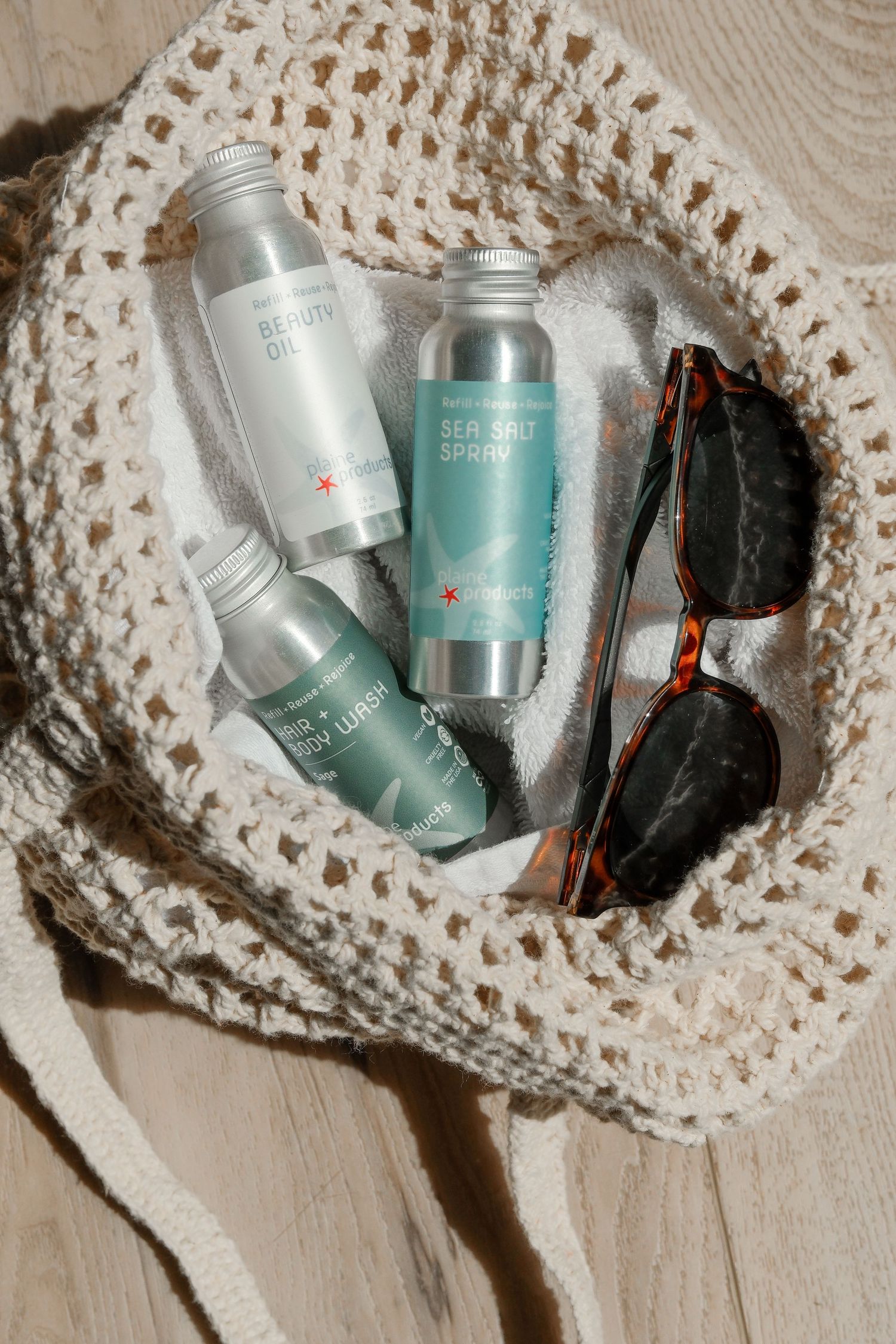 Crochet mesh bag with sea salt spray, beauty oil, hair products, and tortoise shell sunglasses on wooden surface.