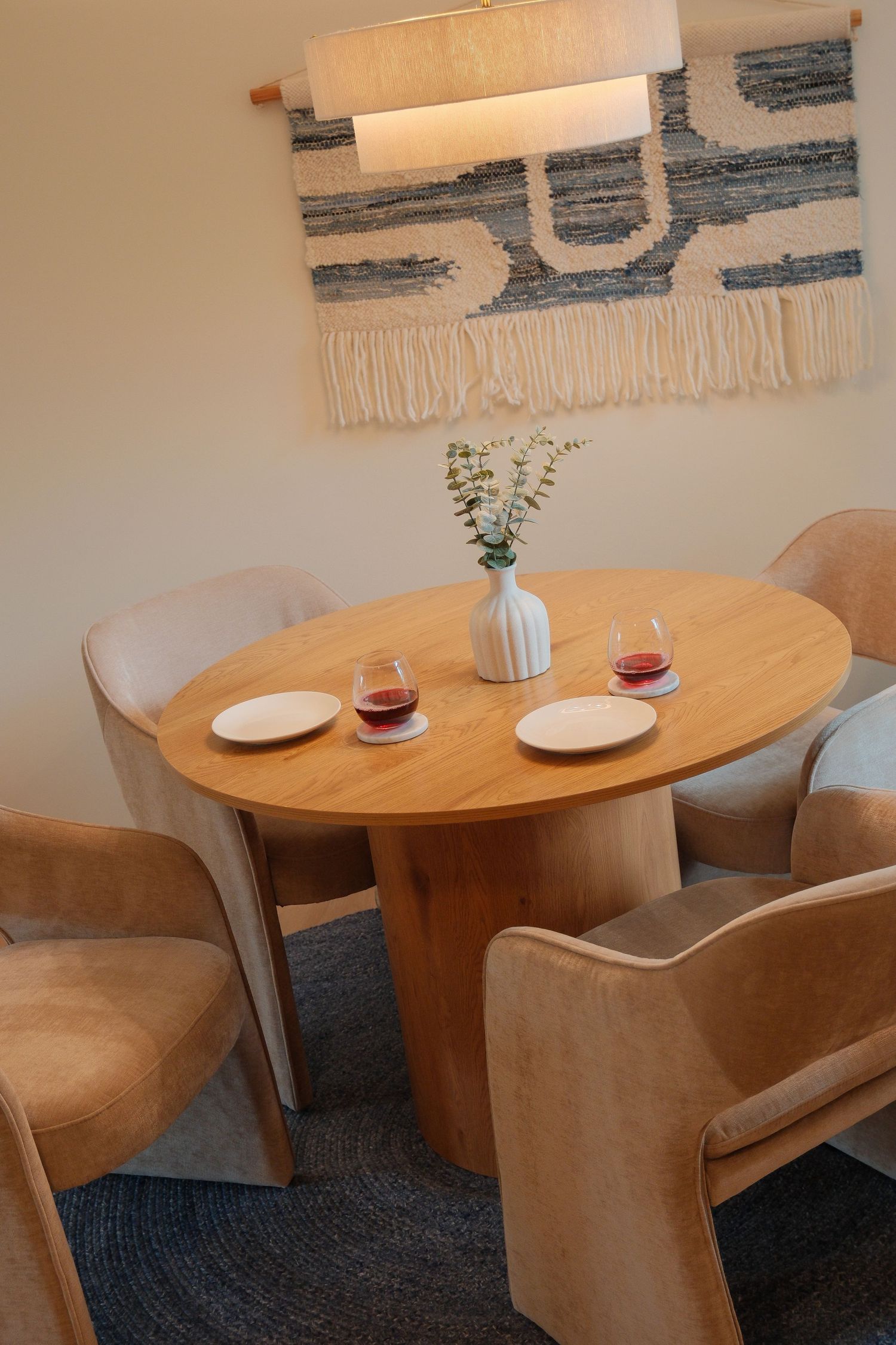 Round wood dining table with tan chairs, white vase, flowers, and wine glasses beneath a woven wall hanging.