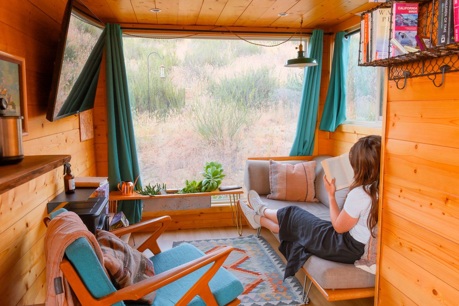 Woman relaxing on a cozy wooden tiny home interior with teal curtains and a scenic desert view through large windows.