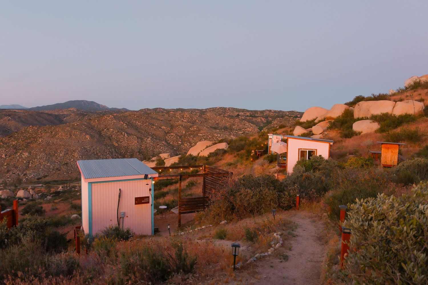 Small white buildings on a rocky hillside at sunset with mountain views and desert scrub vegetation in warm golden light.