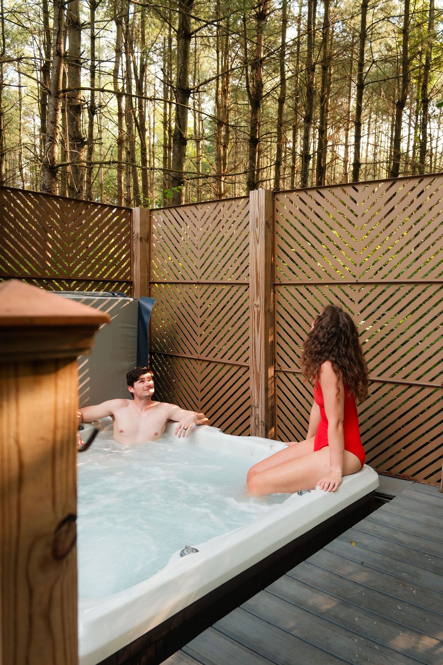 Couple relaxing in an outdoor hot tub on a wooden deck surrounded by tall trees and a decorative fence.