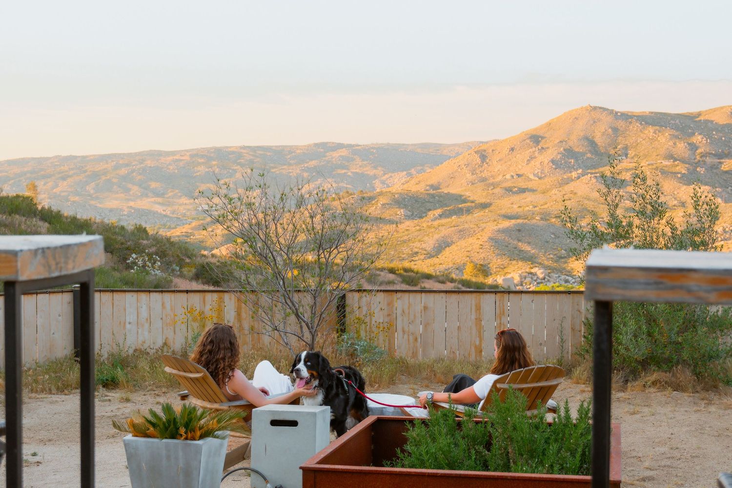 Two women relax on a patio with a dog, enjoying a scenic desert mountain view at golden hour.