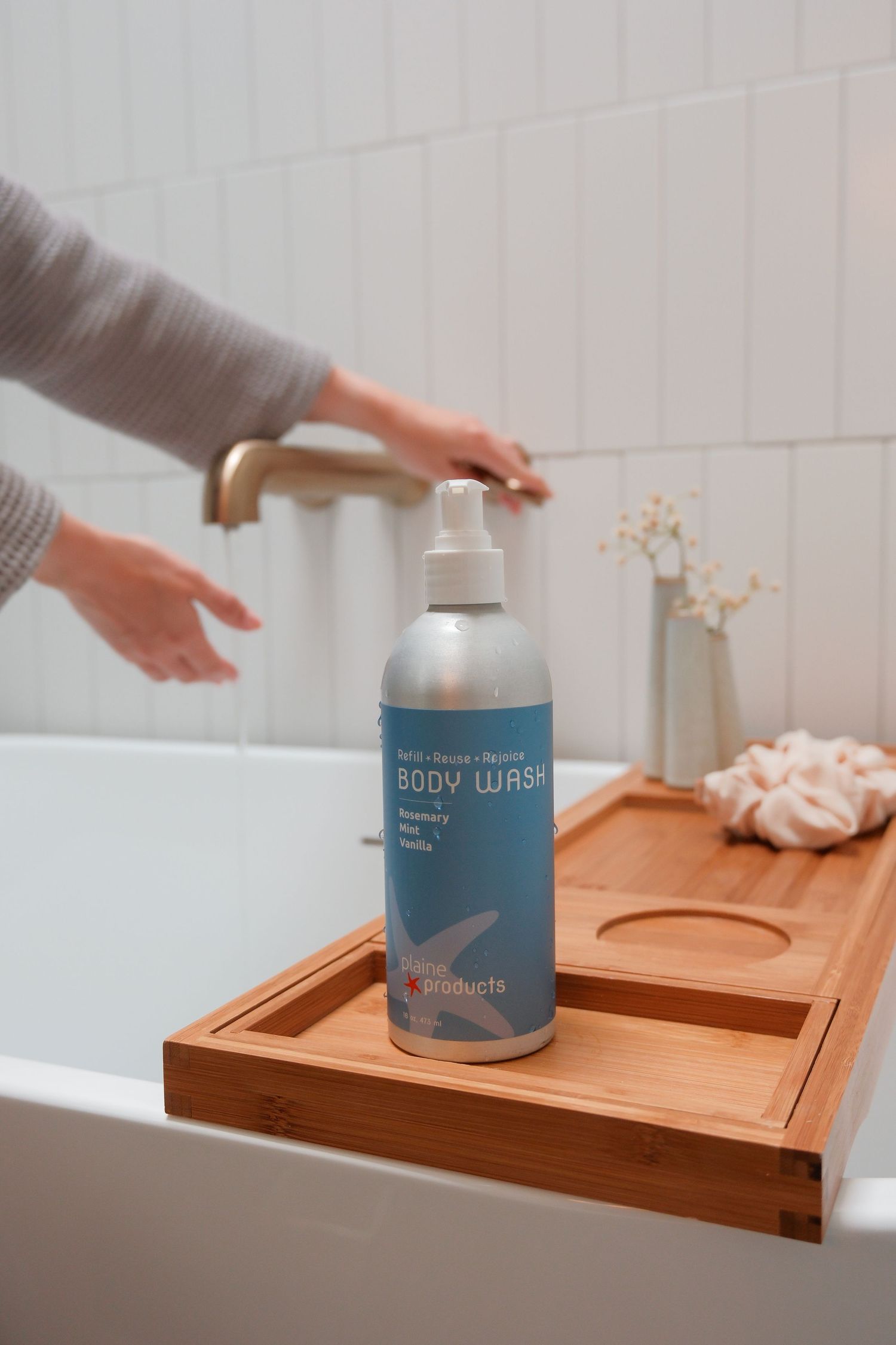 Plain Products Body Wash bottle in teal label displayed on bamboo bath tray near running faucet.