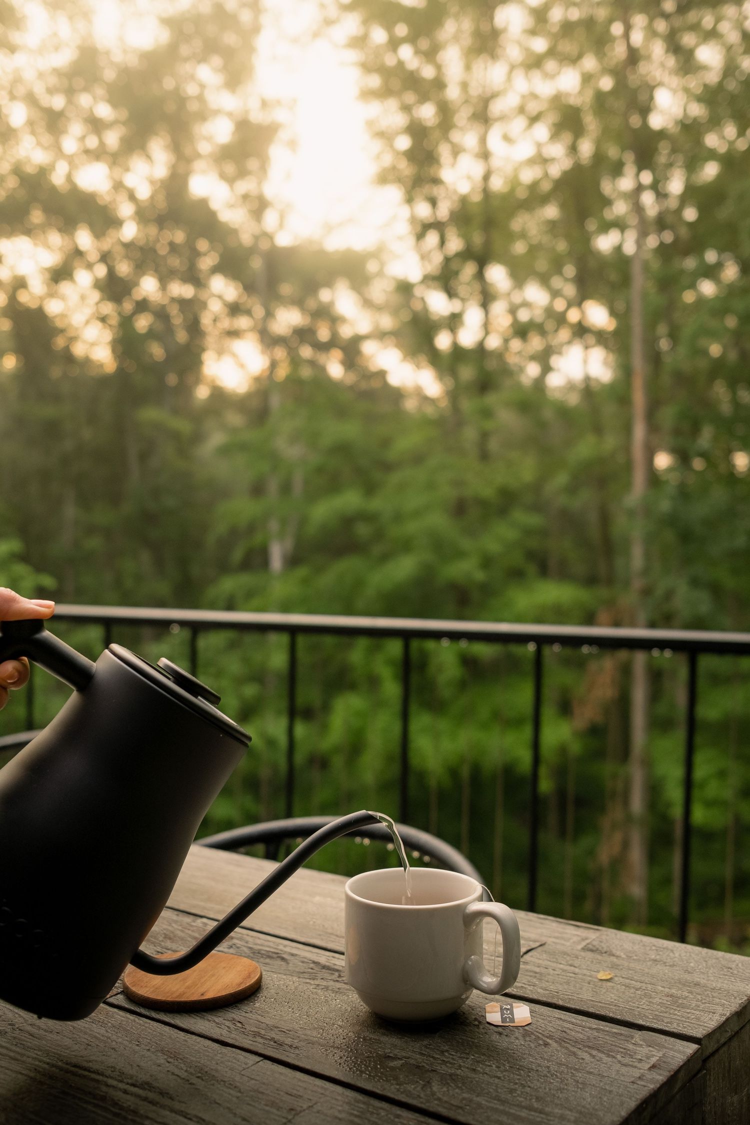 Black kettle pouring hot tea into white cup on wooden deck railing overlooking lush forest at sunset.
