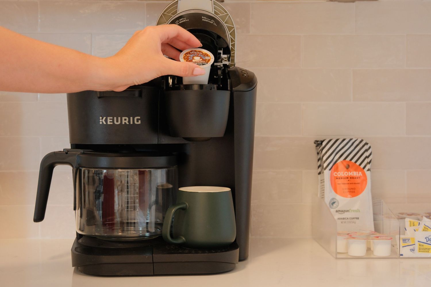 Hand inserting a K-Cup pod into a black Keurig coffee maker with a green mug on the drip tray.
