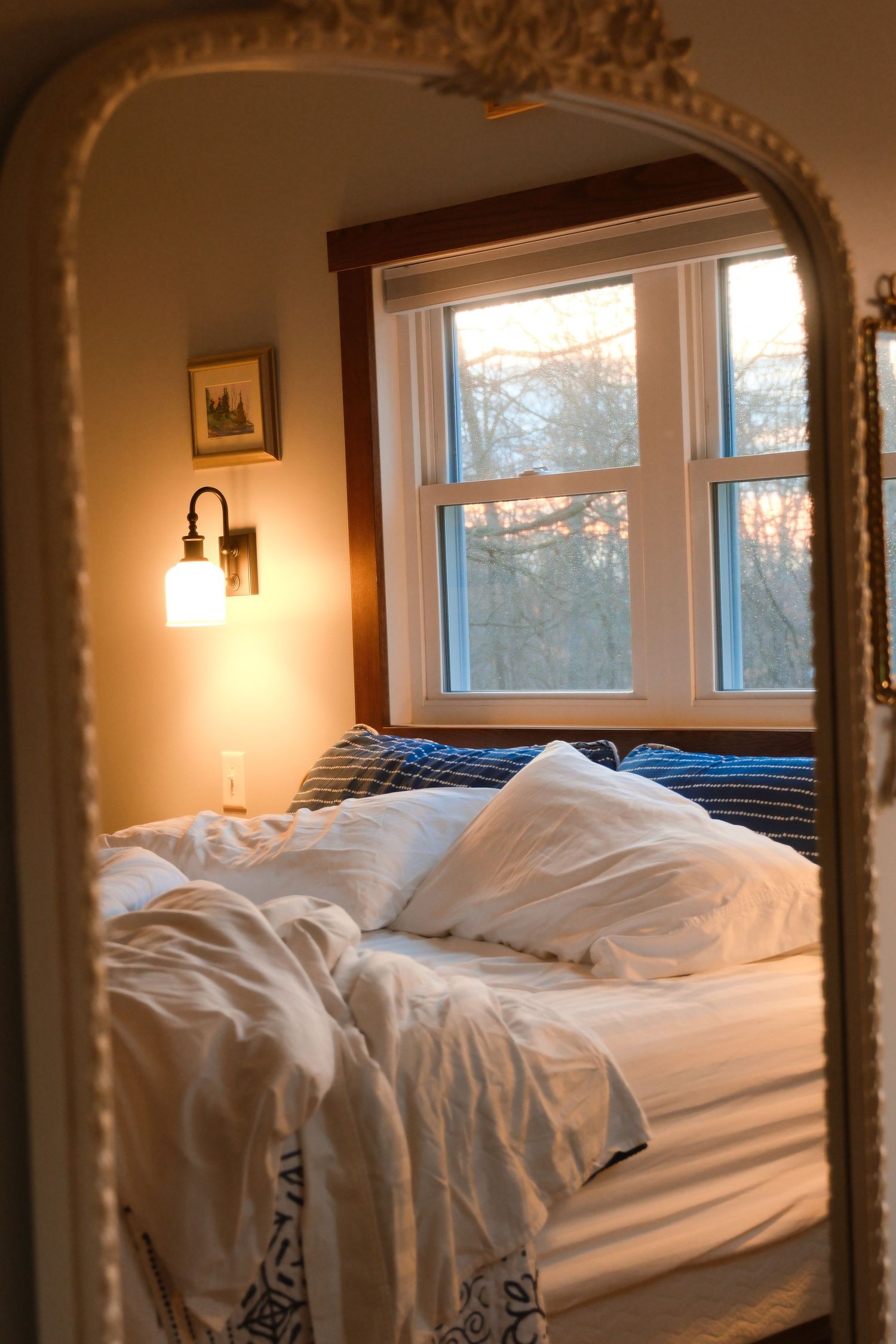 Unmade bed with white linens reflected in an ornate mirror with natural light streaming through windows.