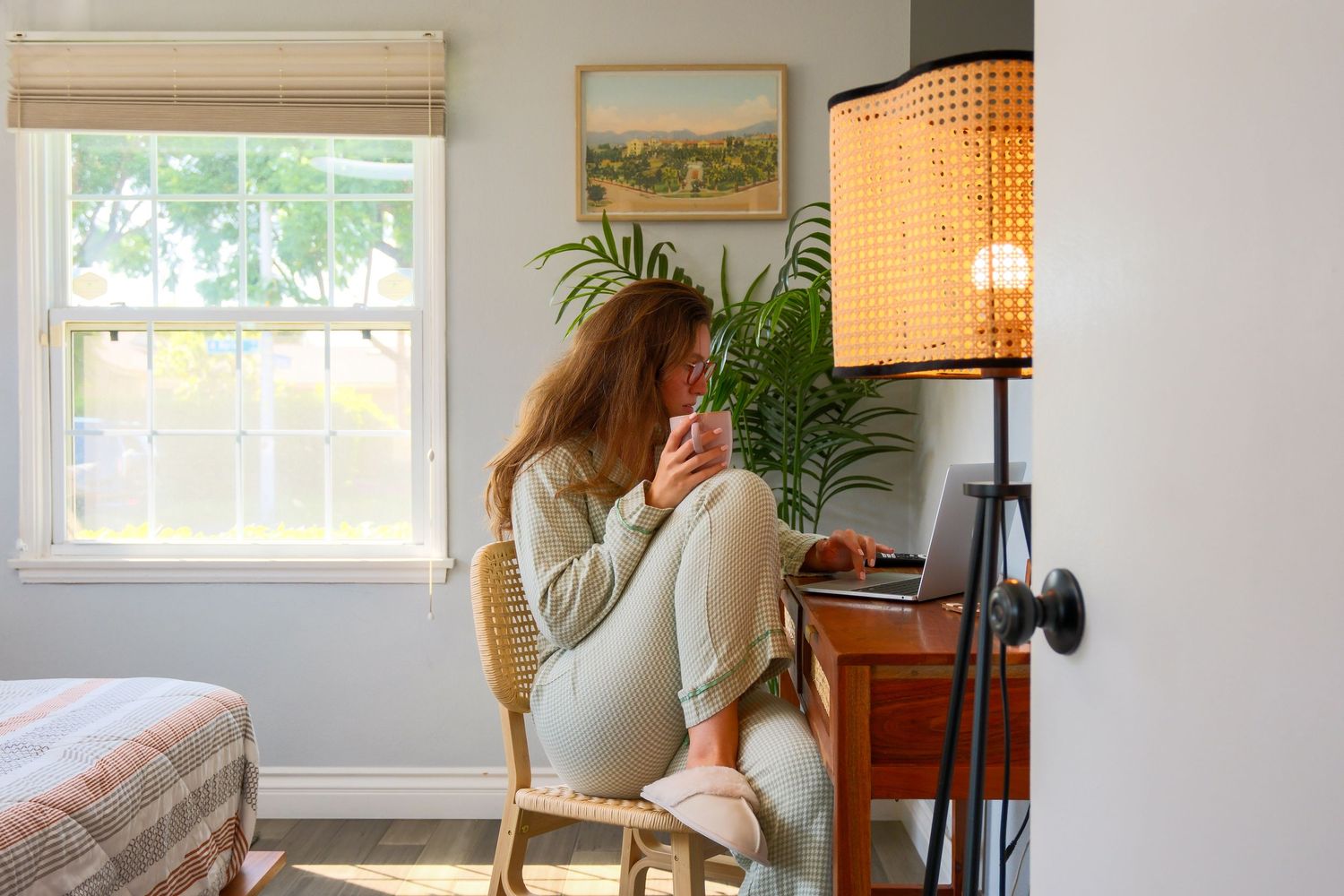 Woman sitting in wicker chair using laptop by window with lamp and houseplant in cozy home office.