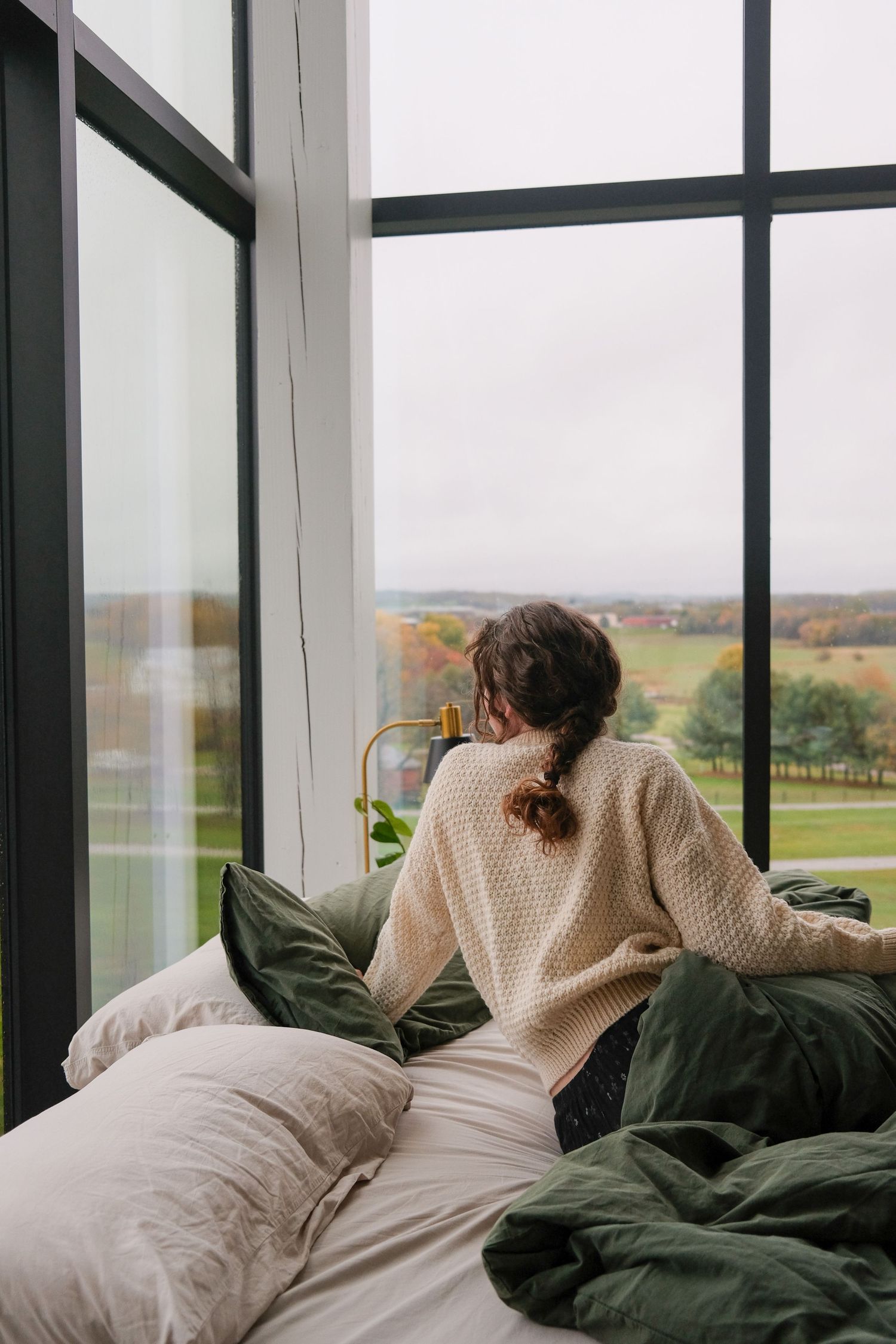 Woman in cream knit sweater sitting on bed with green pillows, gazing out large floor-to-ceiling windows at autumn countryside.