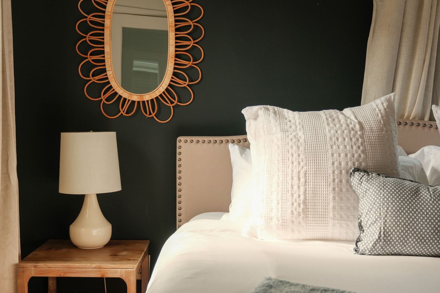 Cozy bedroom with dark green wall, rattan sunburst mirror, white lamp on wooden nightstand, and neutral toned pillows on bed.