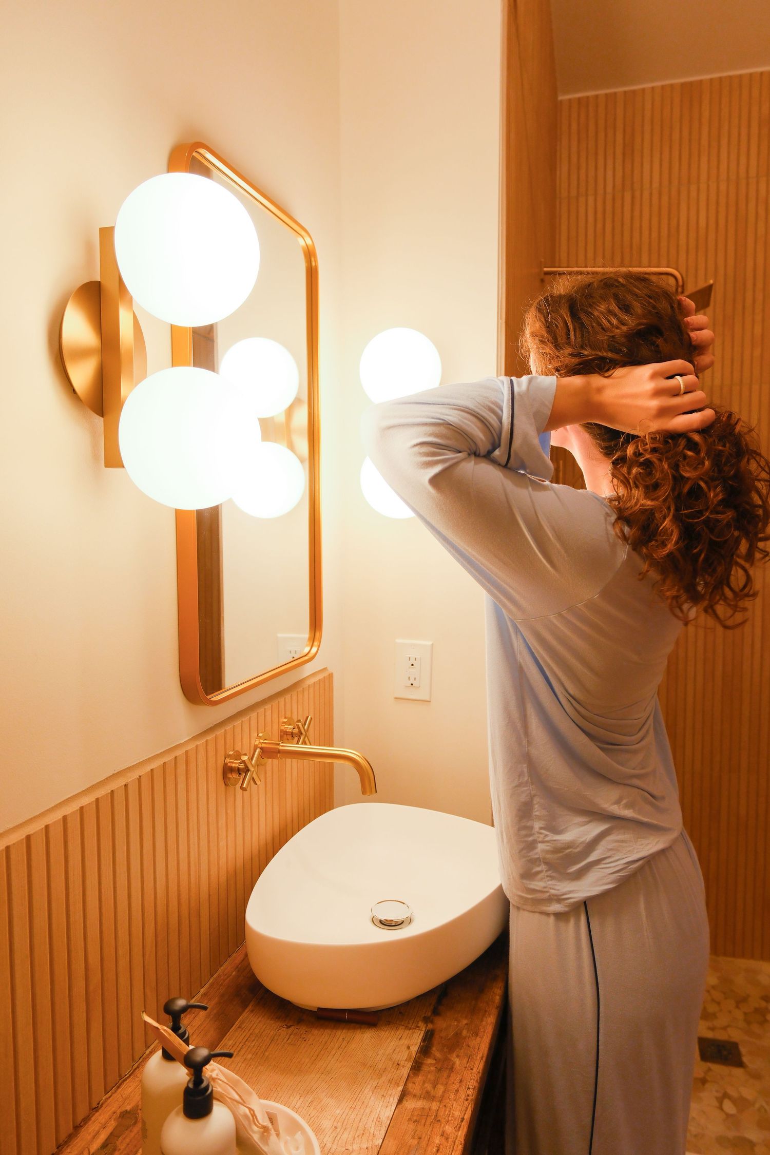 Woman in blue pajamas styling curly hair at a warm-lit bathroom vanity with gold fixtures and globe lights.