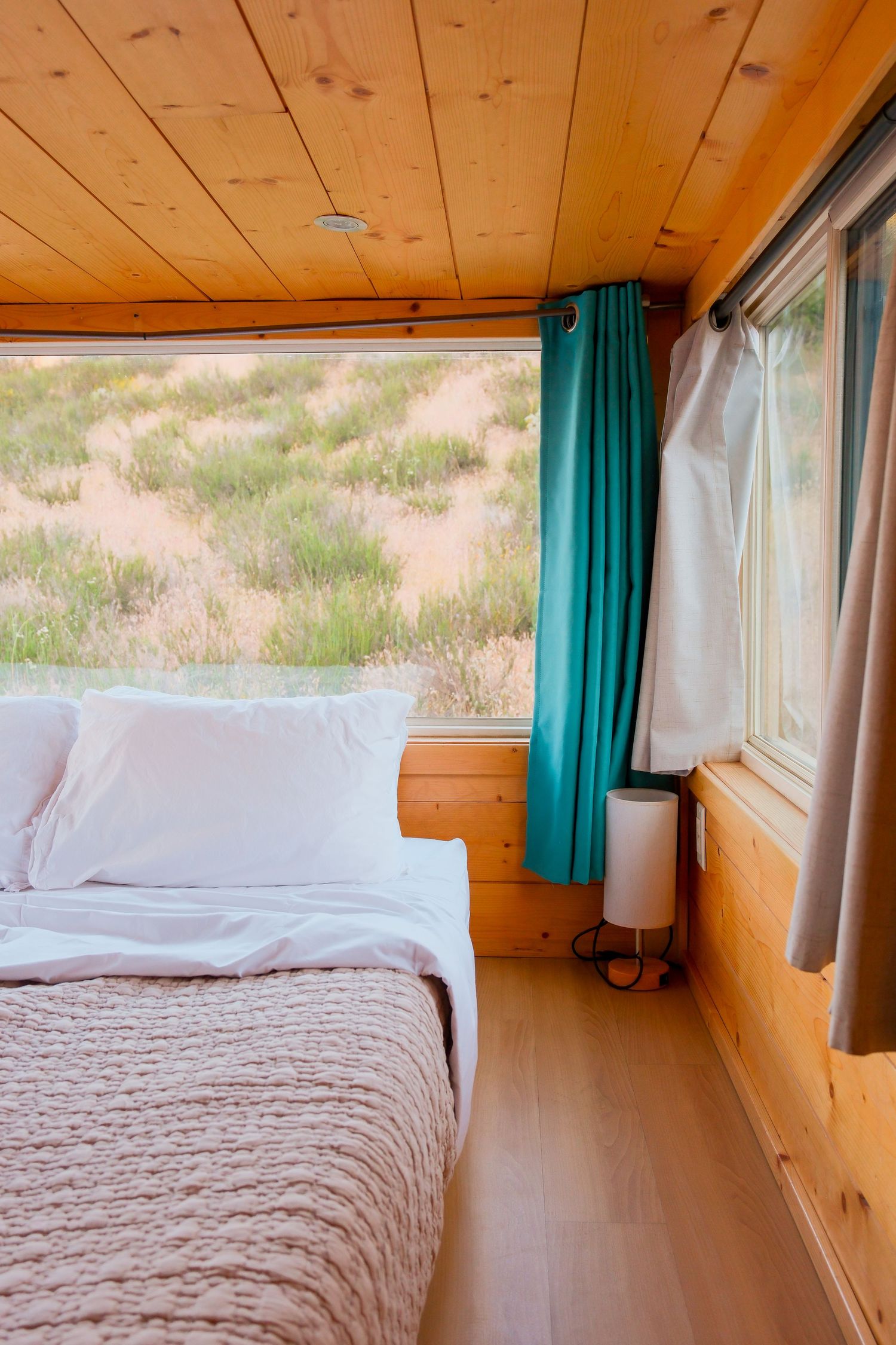 Cozy wooden cabin bedroom with white bedding, teal curtains, and large windows overlooking a scenic desert landscape.