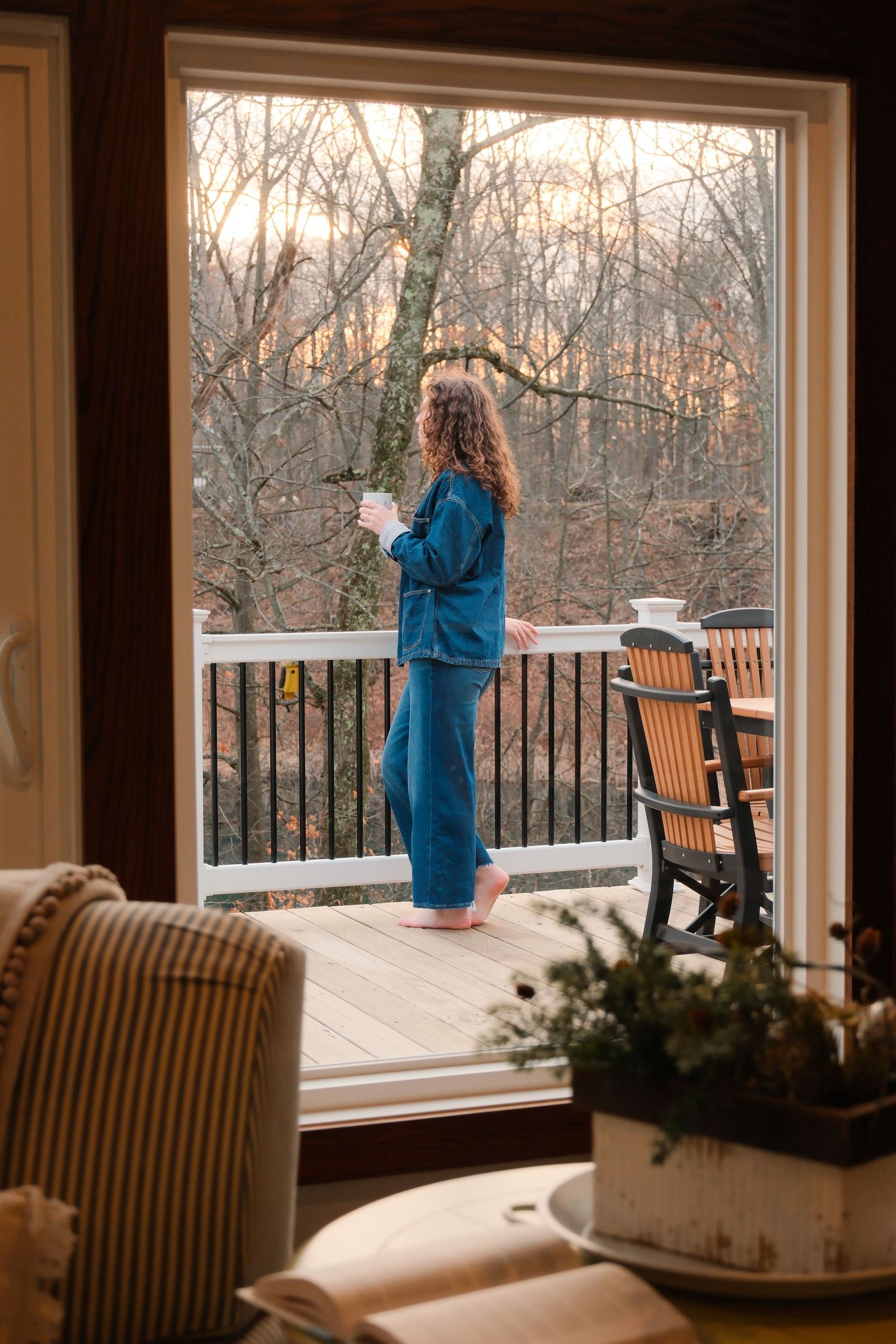 Woman in blue pajamas standing on a wooden deck holding coffee, overlooking bare winter trees at sunset.