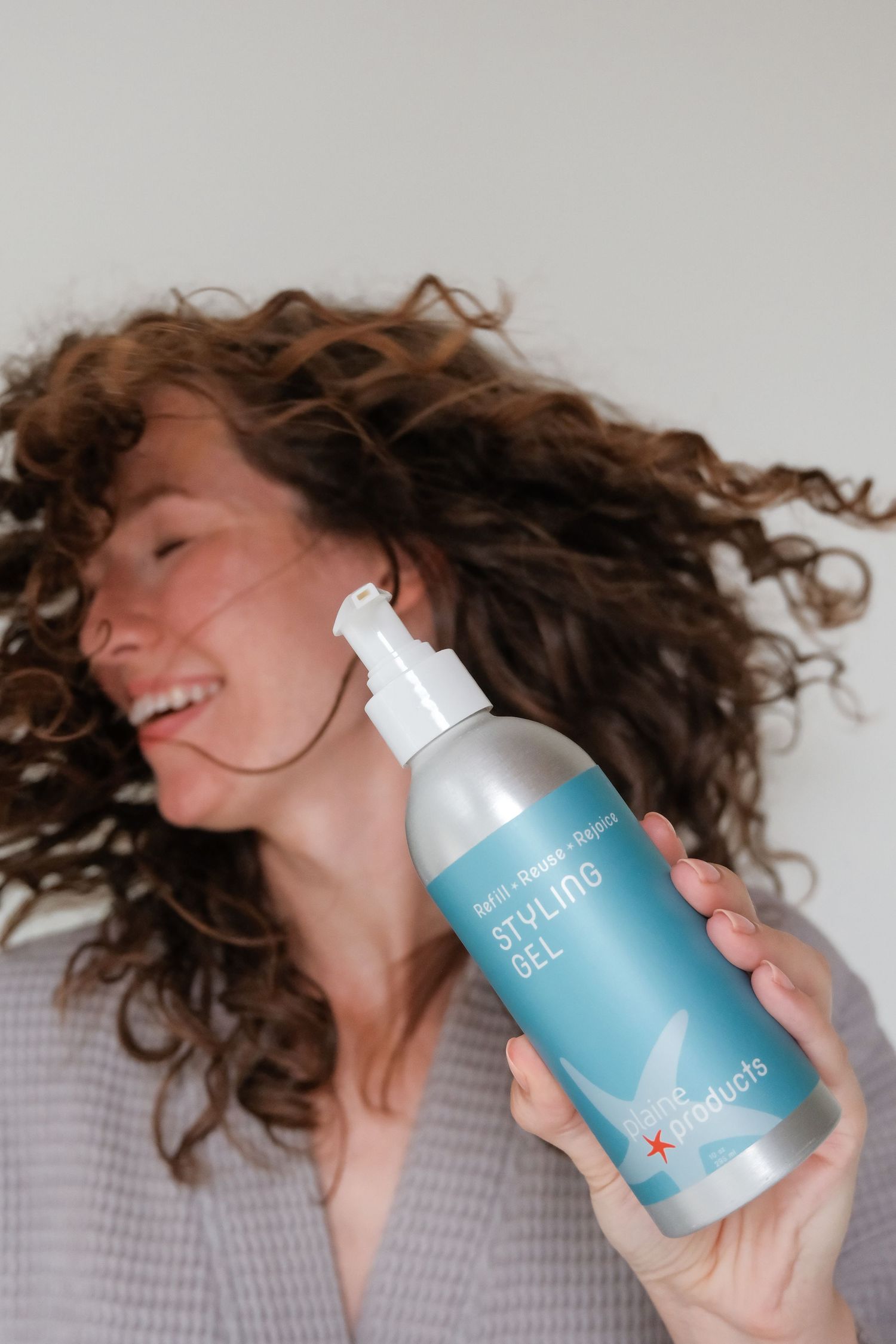 Smiling woman with curly hair holding a pump bottle of styling gel, showcasing voluminous, bouncy curls.