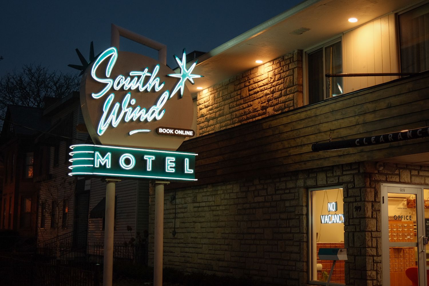Neon sign for South Wind Motel glowing at dusk, mounted on brick building with warm lighting.