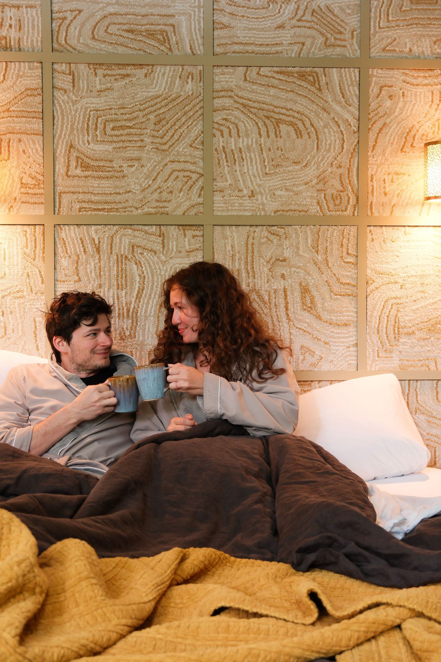 Two people relaxing in bed holding blue mugs, smiling at each other under cozy blankets with warm ambient lighting.