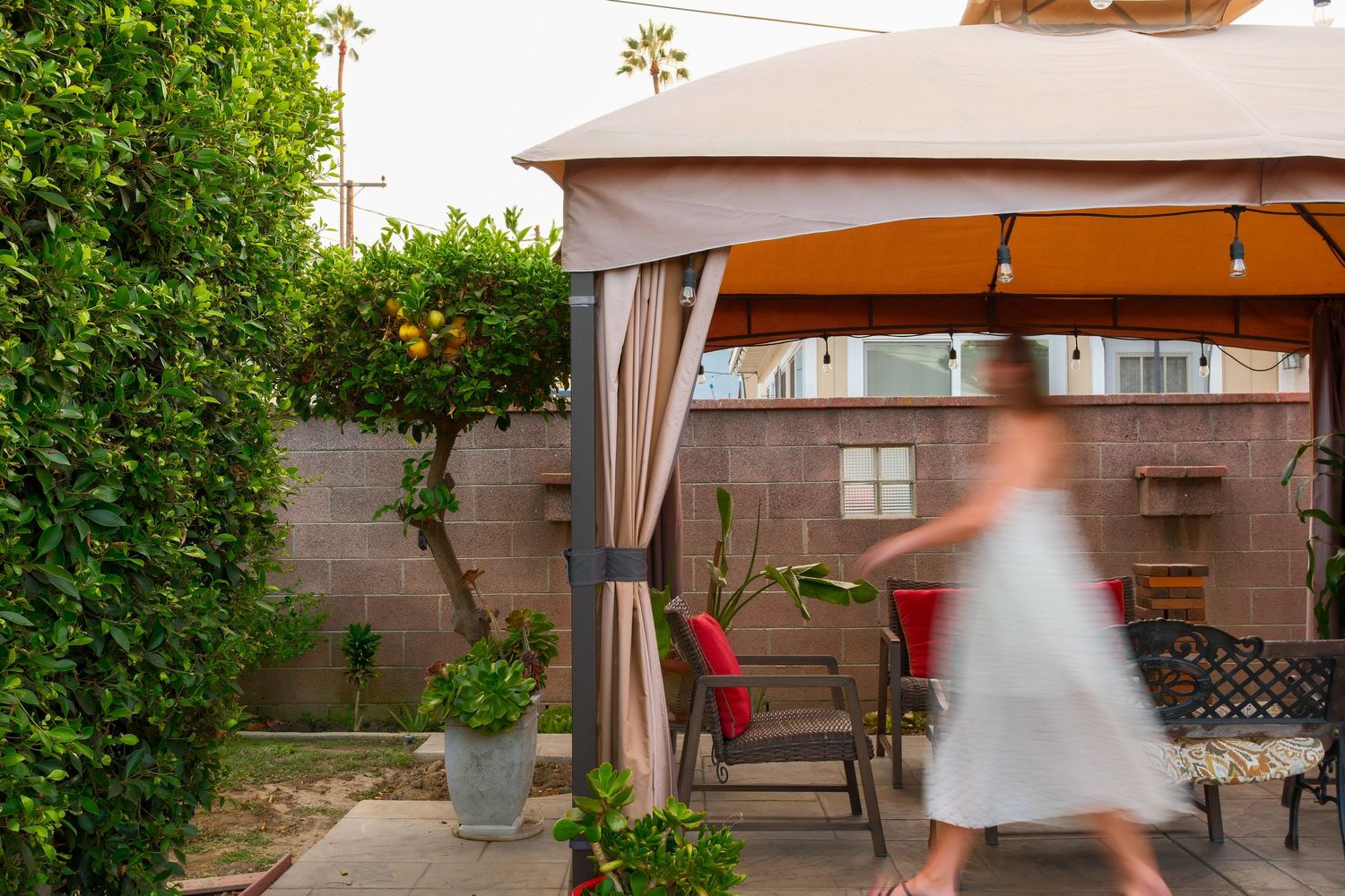 A woman in a white dress walks beneath a beige gazebo in a lush backyard patio with string lights and garden furniture.