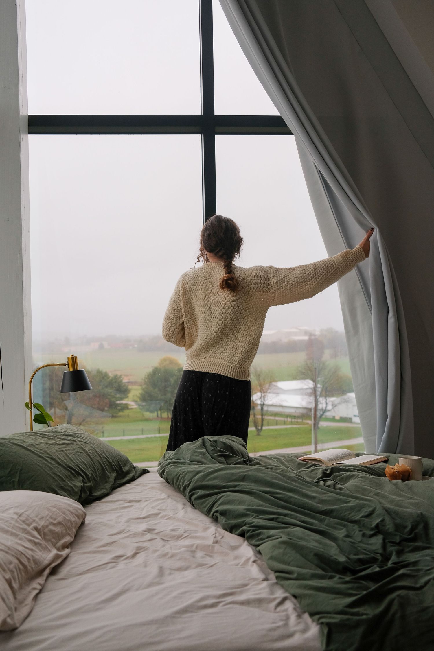Person stretching in bed by large windows overlooking a scenic rural landscape on a cloudy morning.