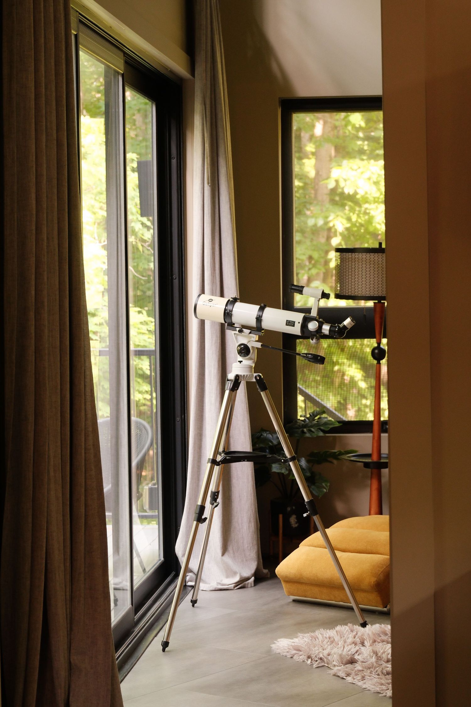 A white telescope on a tripod stands by large windows with forest views in a modern, warmly decorated living room.