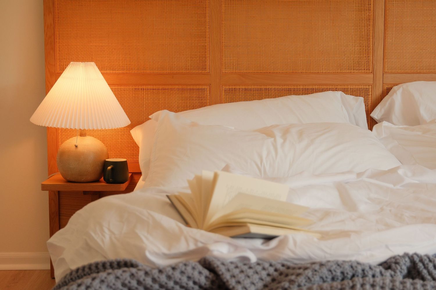 Cozy bedroom with white bedding, open book, glowing table lamp on nightstand, and gray knit blanket.