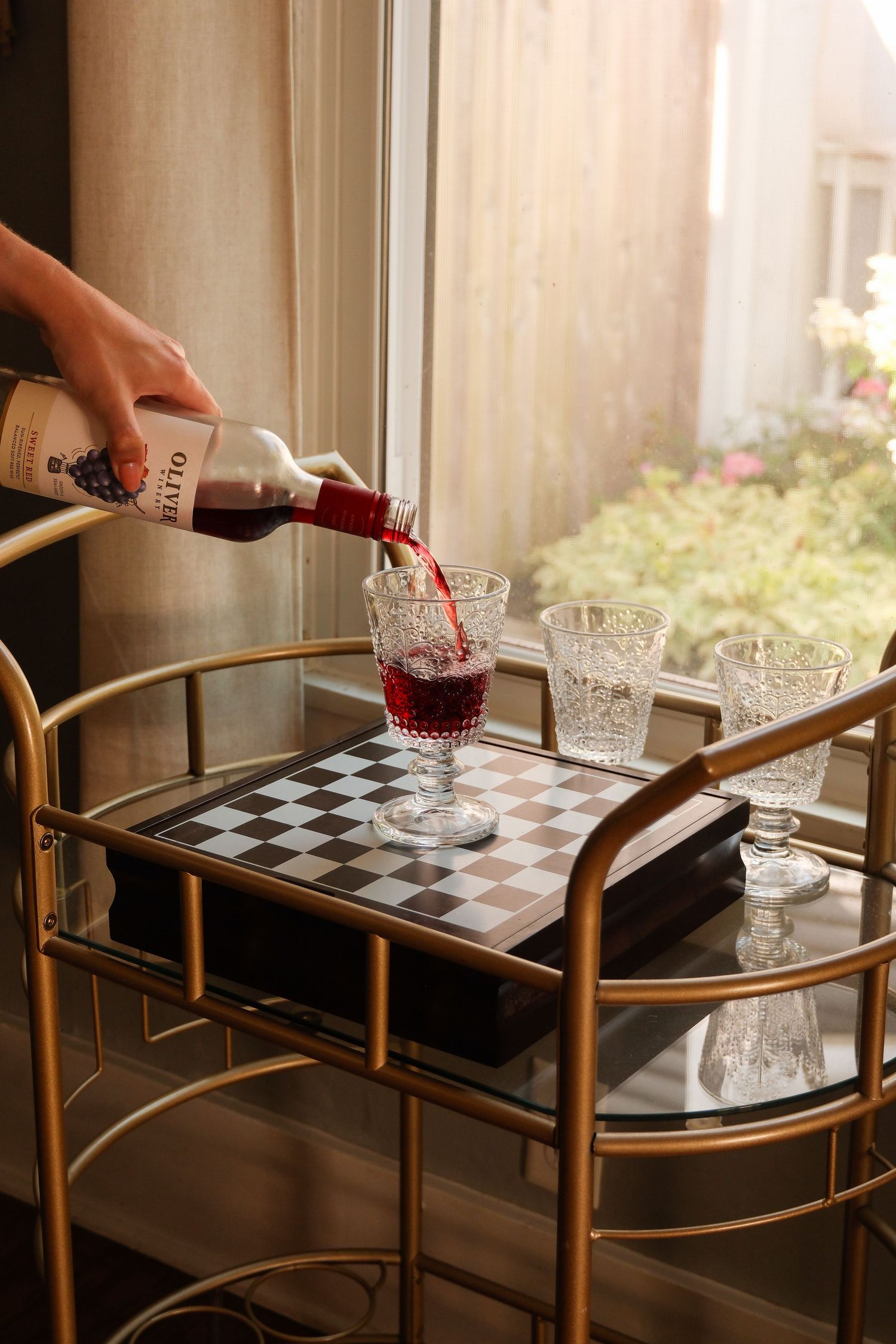 Hand pouring red wine into crystal glasses on a gold bar cart with chess board near a sunlit window.