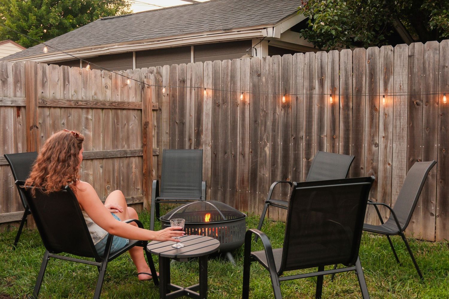 Woman relaxing in backyard patio with fire pit, string lights, and black outdoor chairs near wooden fence.