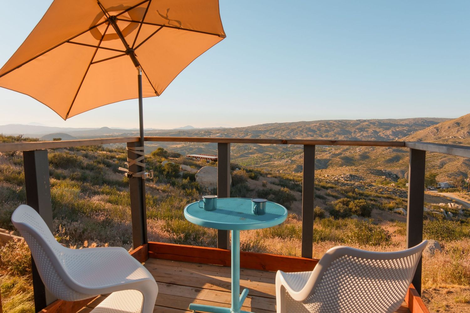 Outdoor deck with teal table, white chairs, and orange umbrella overlooking a scenic desert valley landscape.