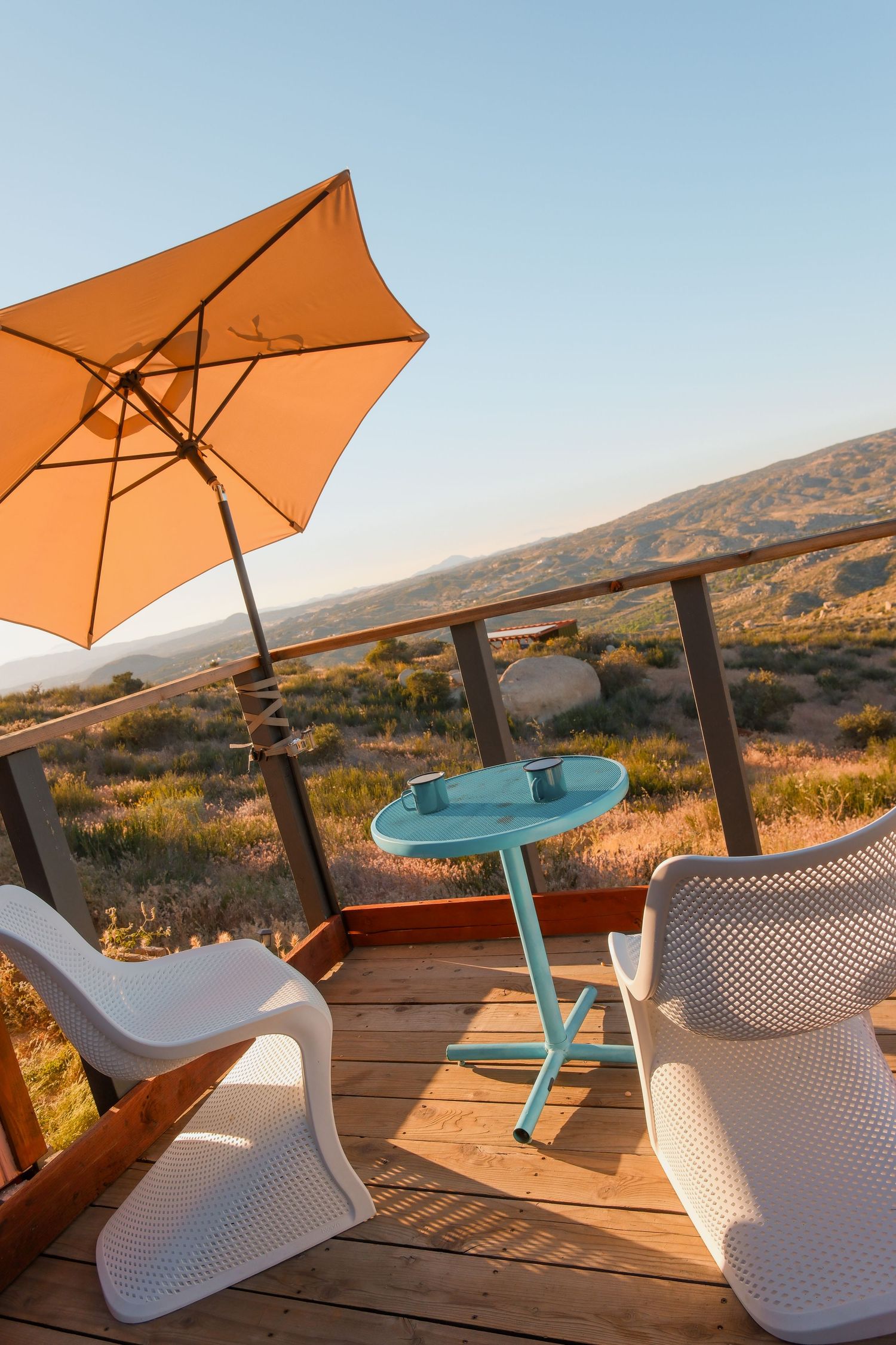 Outdoor deck with white chairs, teal table, and orange umbrella overlooking a scenic desert landscape at golden hour.