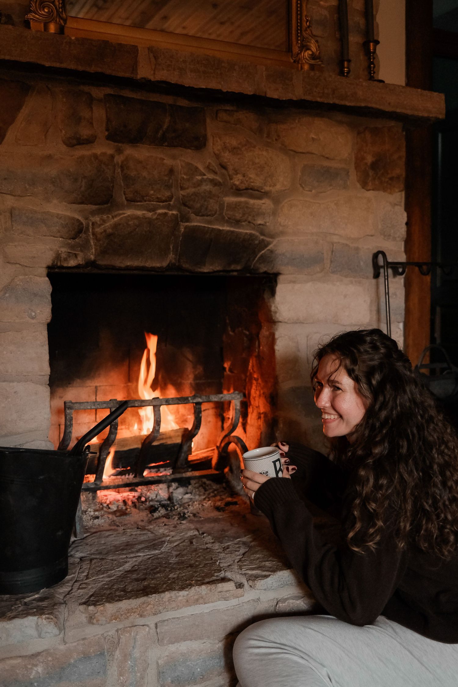 Cozy stone fireplace with glowing flames and someone enjoying a warm drink.