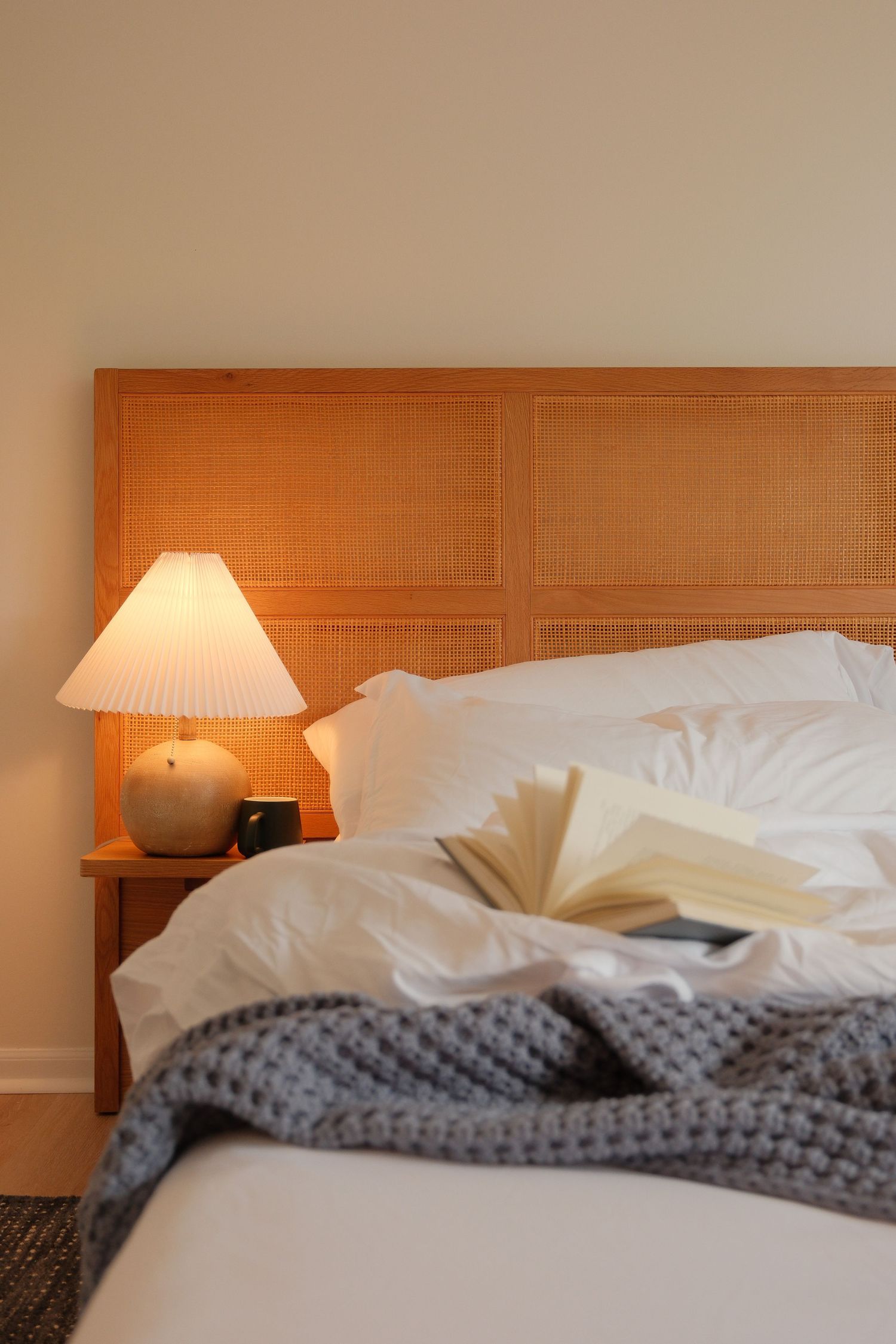Cozy bedroom with wooden headboard, glowing bedside lamp, open book, and gray knit blanket on white bedding.