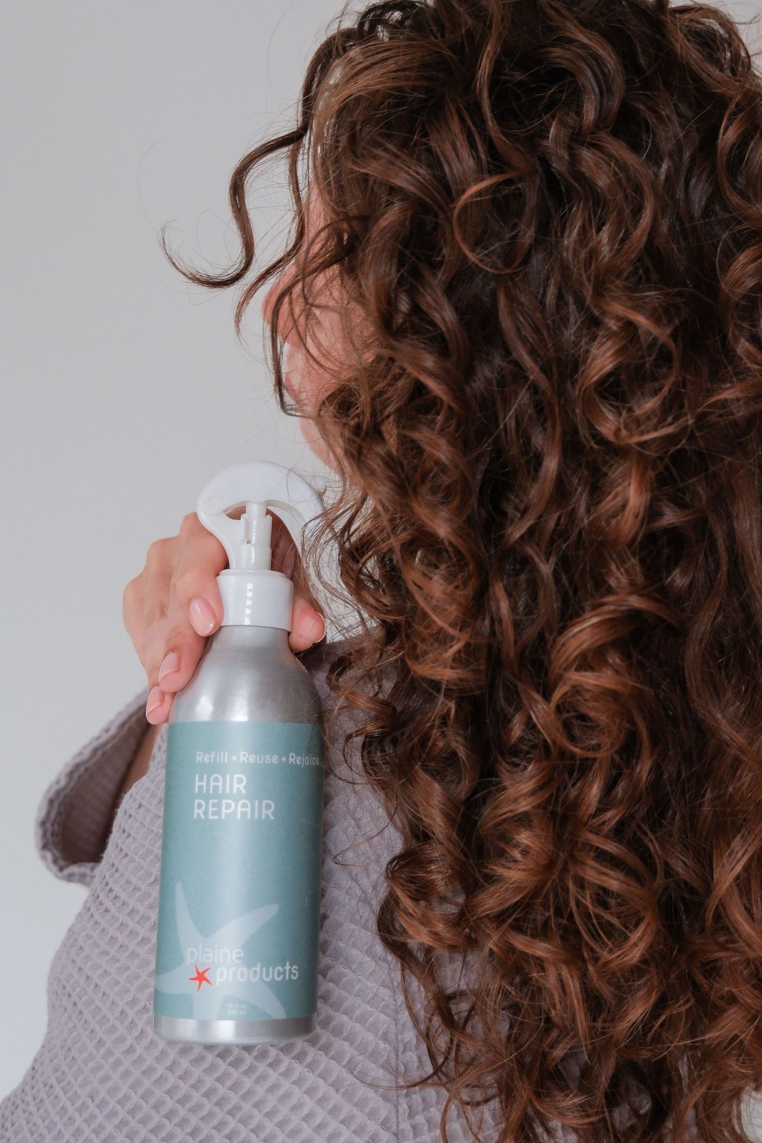Woman with long curly brown hair holding a Hair Repair spray bottle by gz products.