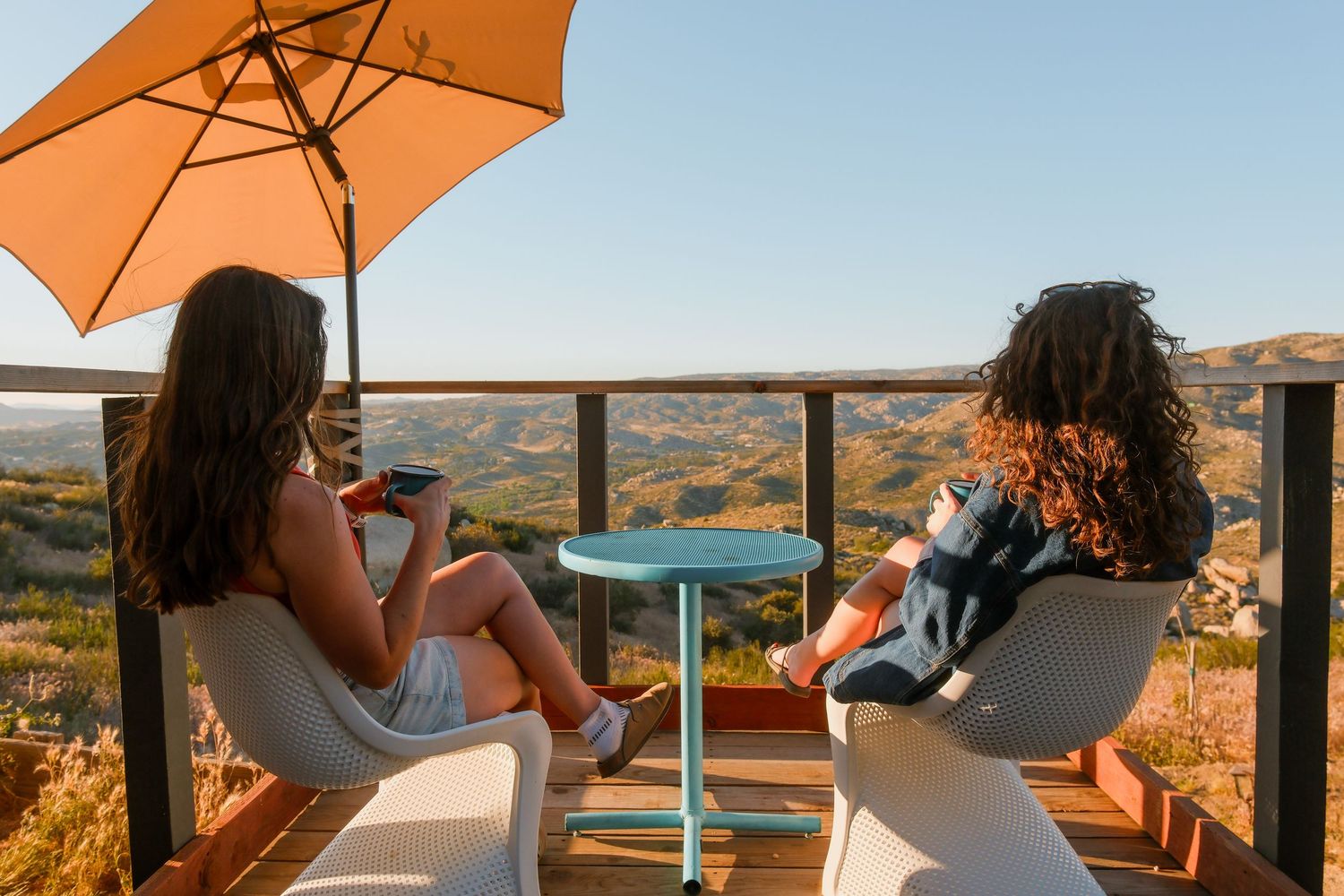 Two women sit in white chairs on a scenic overlook balcony with an orange umbrella and teal table.