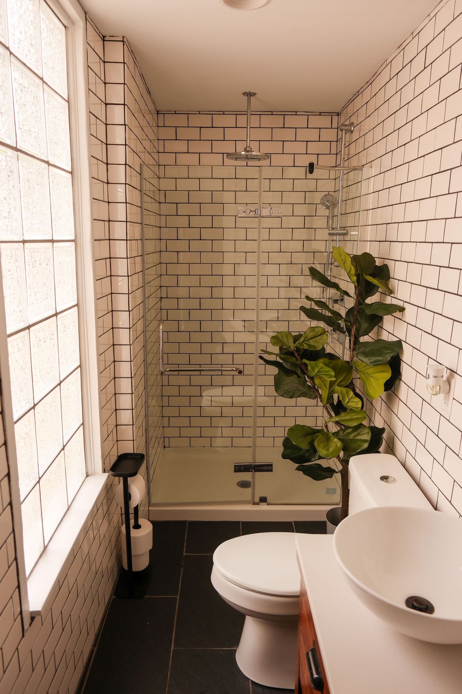 Modern bathroom with white subway tiles, glass shower, vessel sink, dark floor, and fiddle leaf fig plant.