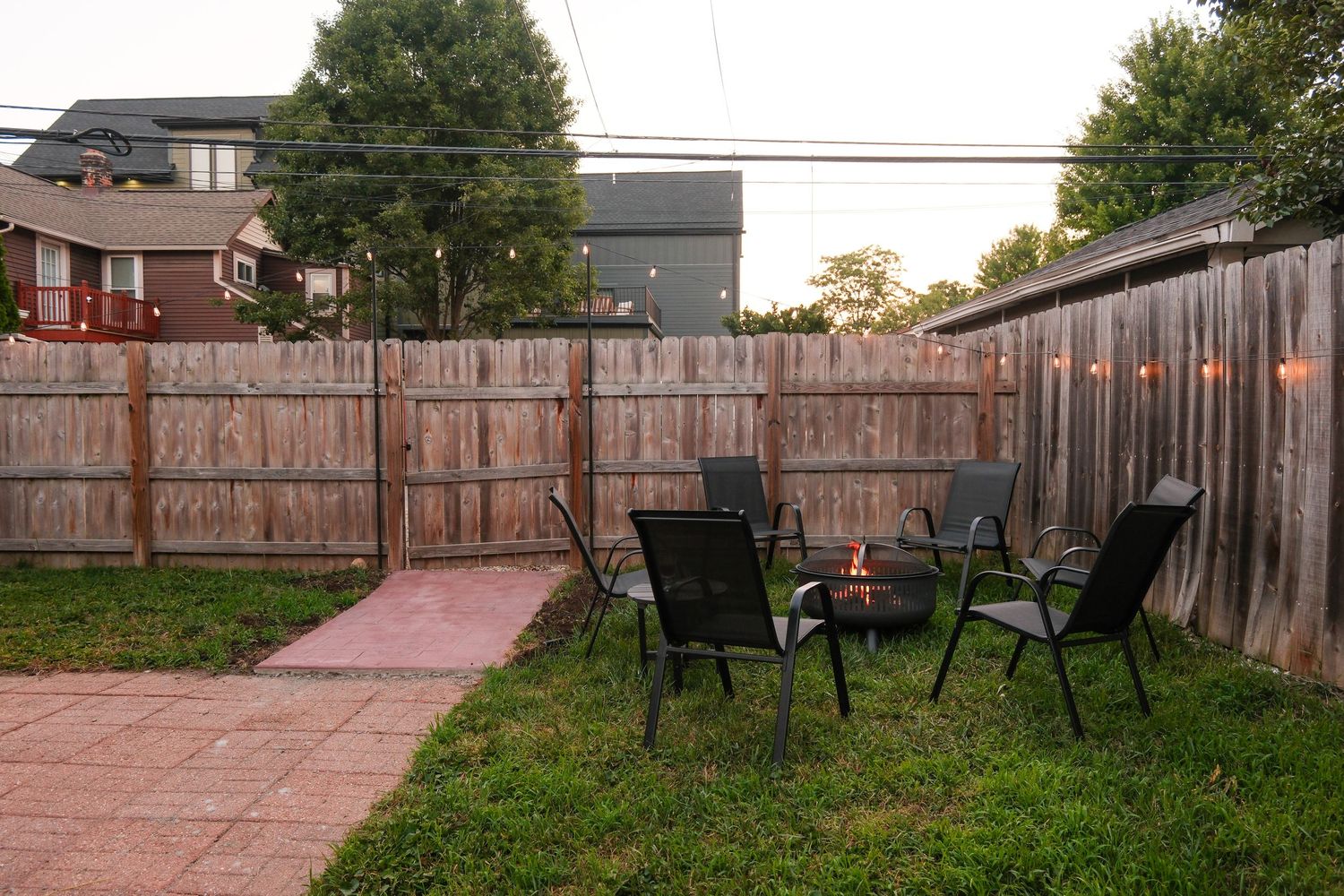 Cozy backyard patio with black chairs around a fire pit, surrounded by a wooden privacy fence and string lights at dusk.