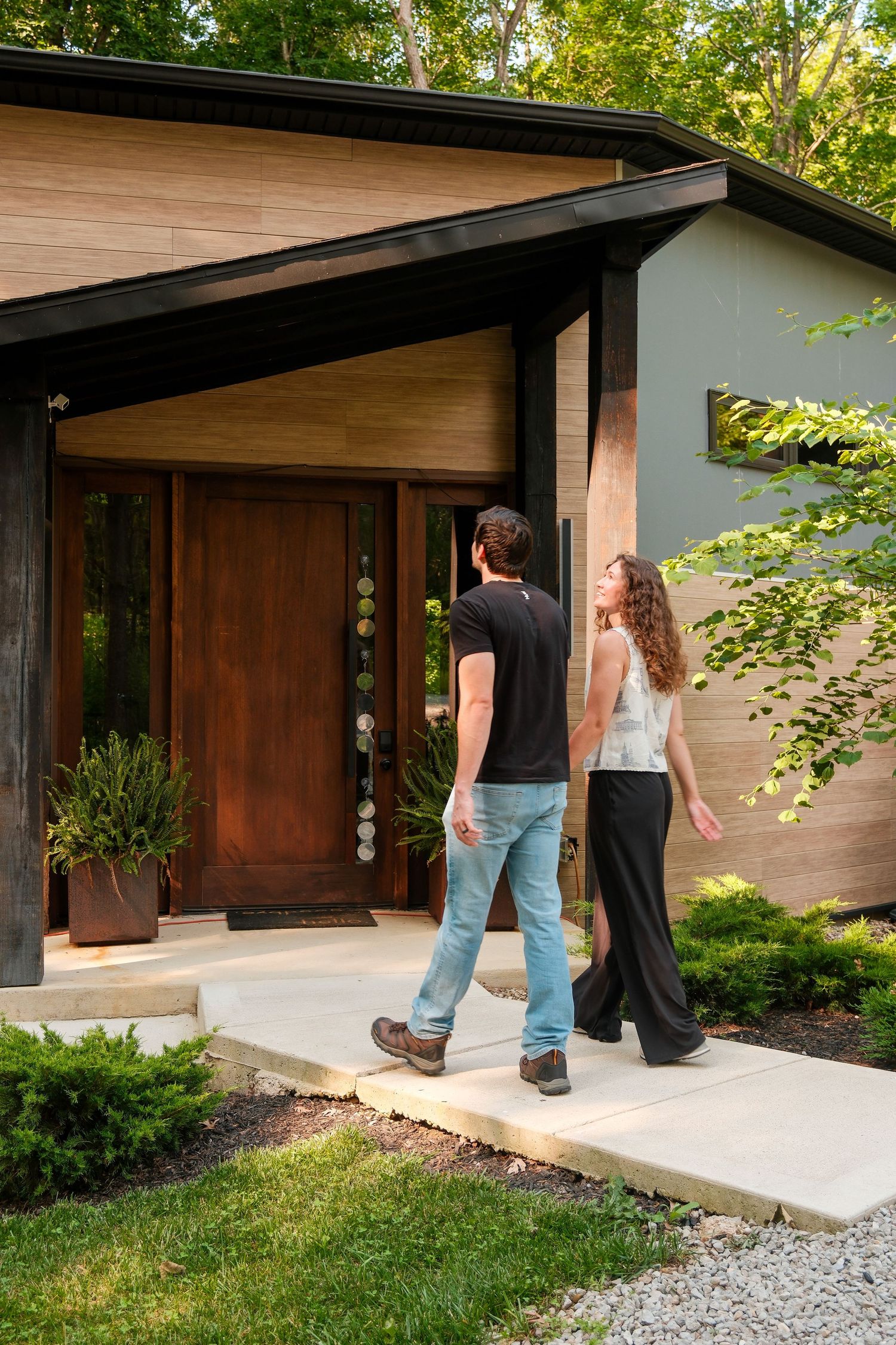 Couple walking toward modern mid-century home entrance with wood door, lush greenery, and clean architectural lines.