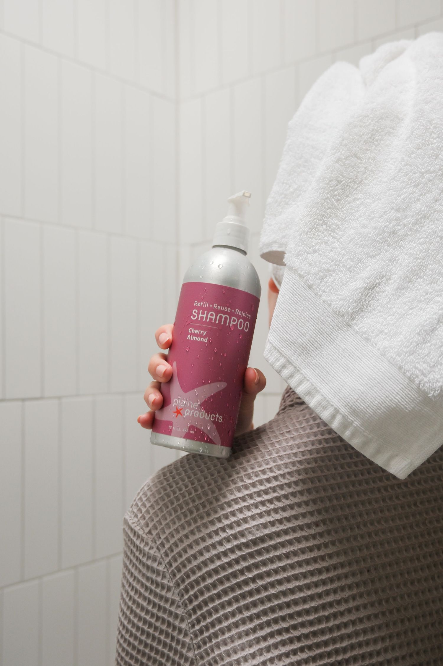 Person in white towel and waffle robe holding a pink cherry shampoo bottle in bathroom.