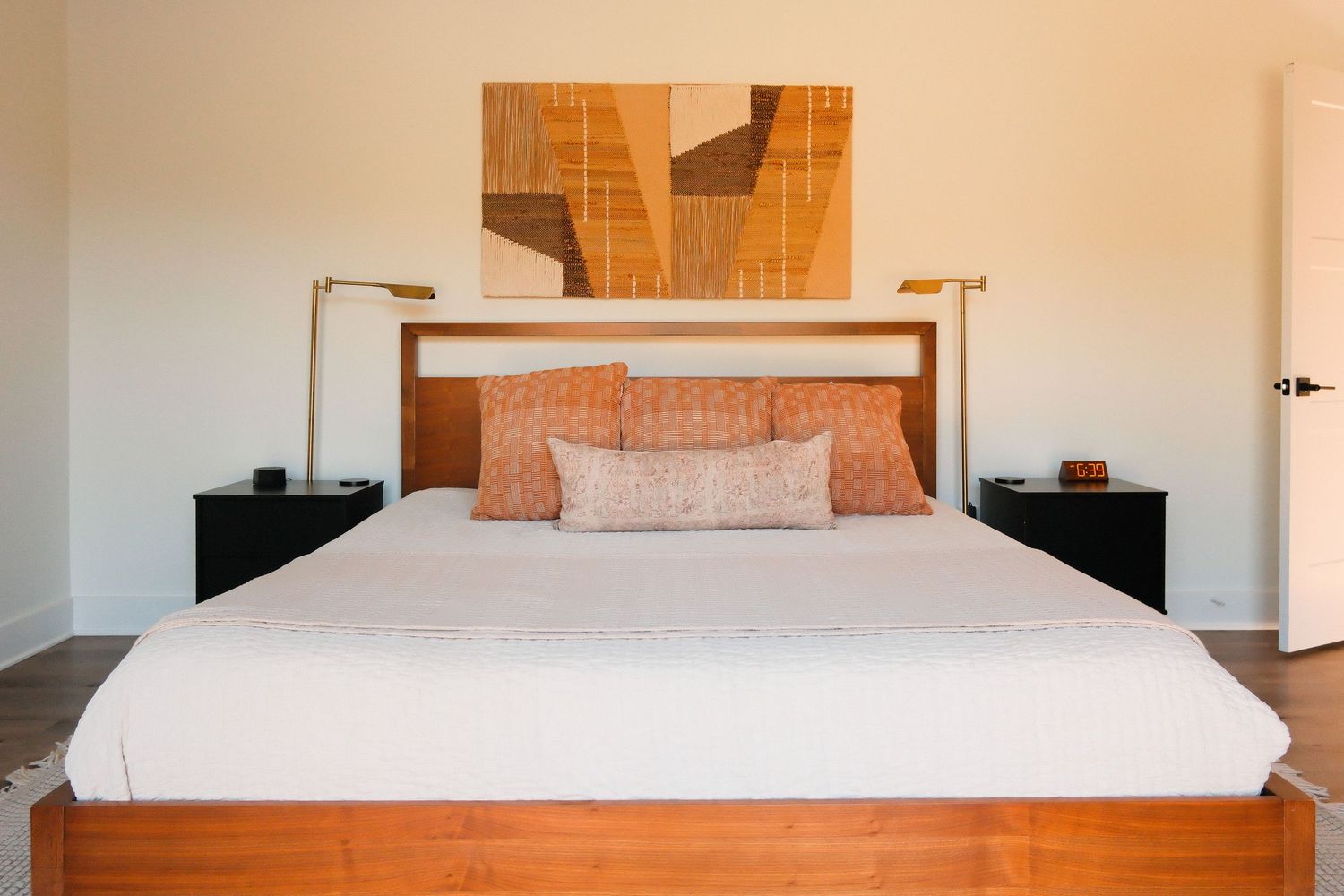 Modern bedroom with wooden bed frame, terracotta pillows, black nightstands, and abstract wall art above.