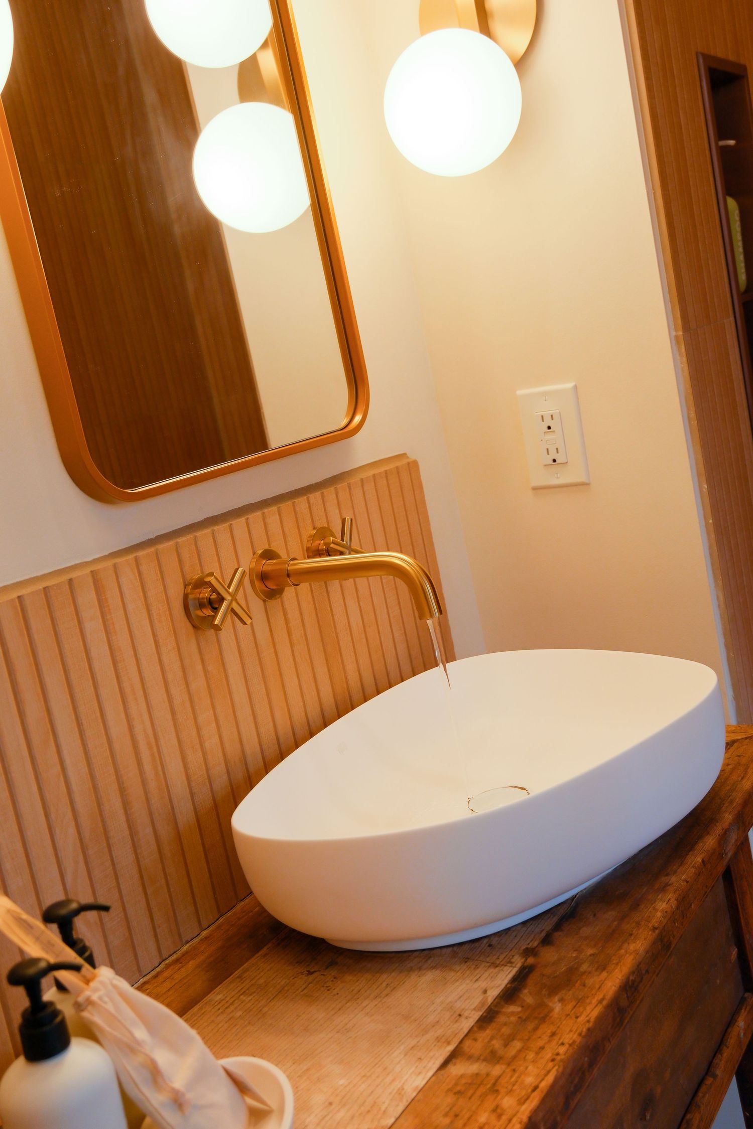 Modern bathroom vessel sink with gold wall-mounted faucet on rustic wood vanity with globe lights and oval mirror.