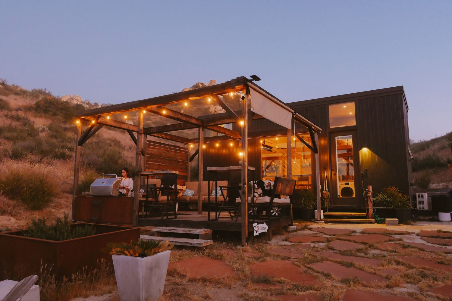 Modern black cabin with string lights on pergola at dusk in desert landscape.