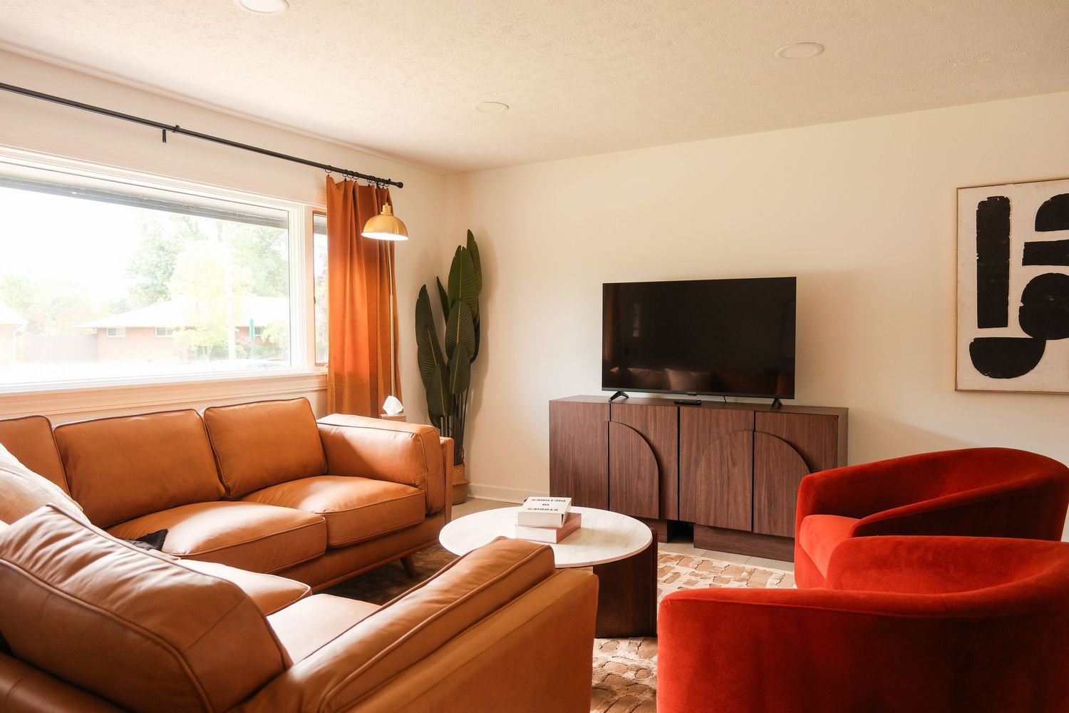 Modern living room with tan leather sofa, red velvet chair, wood TV console, and warm orange curtains.