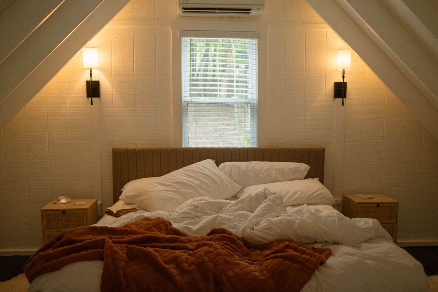 Cozy attic bedroom with white bedding, brown throw, and wall sconces beside a window.