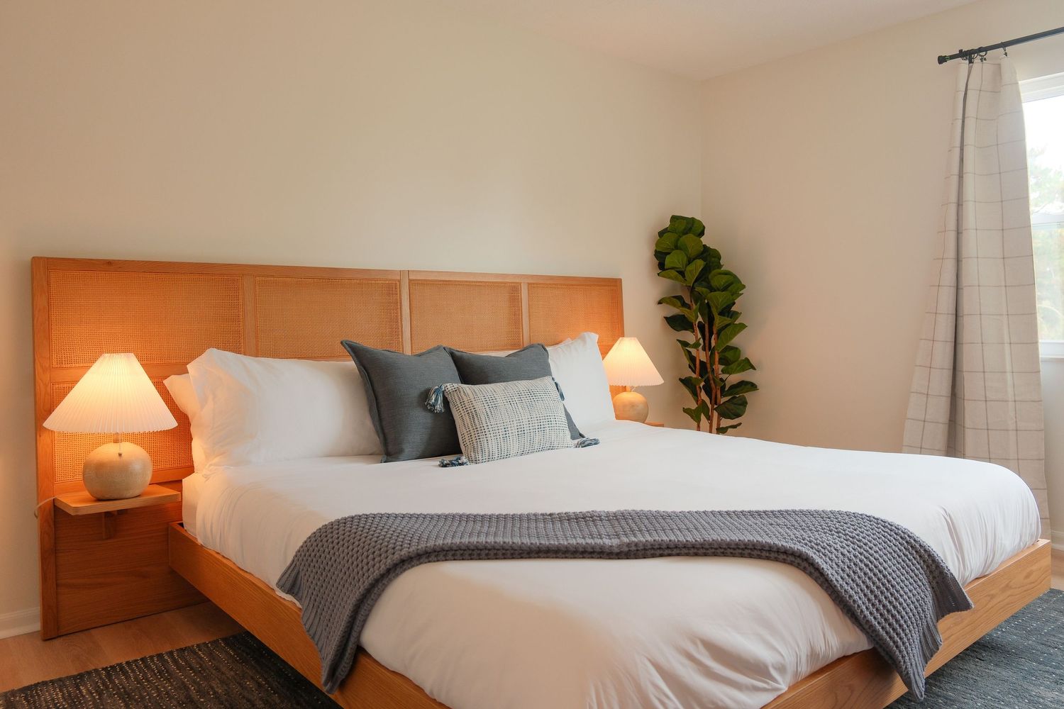 Modern bedroom featuring a king bed with white linens, gray throw, wooden headboard, and a fiddle leaf fig plant.