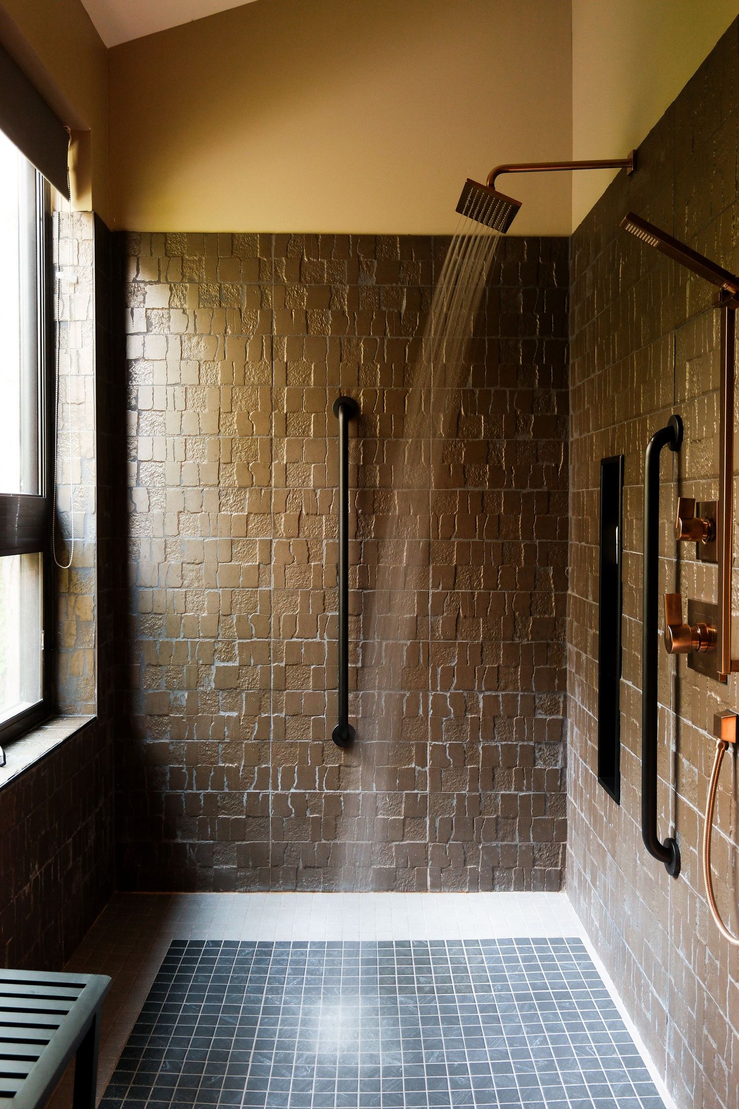 Luxurious walk-in shower with dark textured tiles, copper fixtures, rainfall showerhead, and dark floor tiles in a modern bathroom.