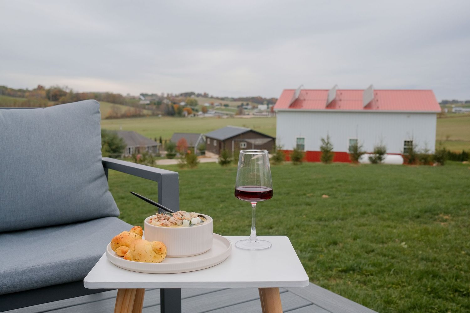 A glass of red wine and cheese board on a white table beside a patio chair overlooking a scenic farm landscape.