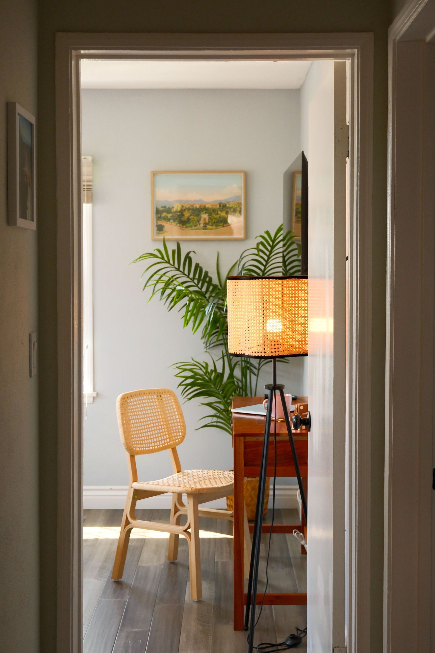 Cozy reading nook with rattan chair, floor lamp, wooden dresser, tropical plant, and framed landscape art on pale blue wall.