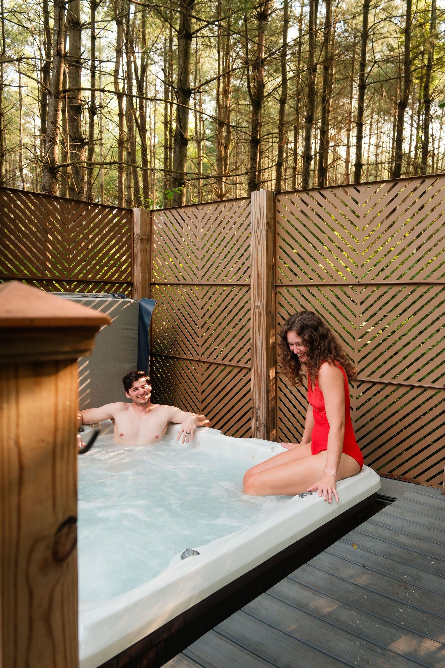 Couple relaxing in an outdoor hot tub on a wooden deck surrounded by tall trees and decorative privacy fencing.