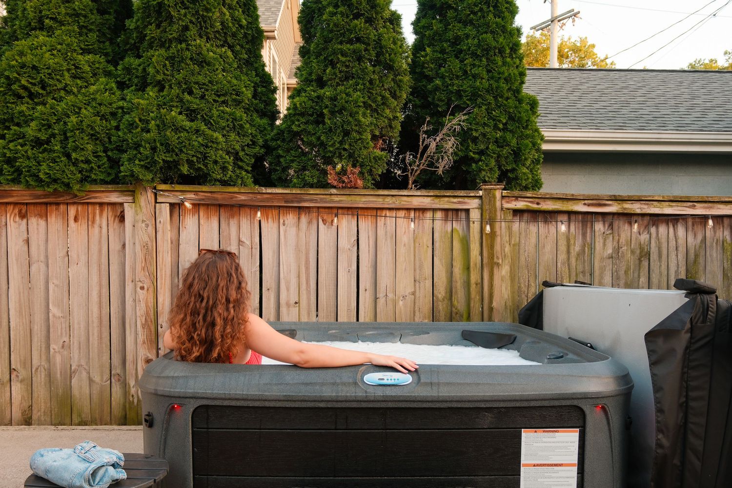 Woman with curly hair relaxing in an outdoor hot tub in a backyard surrounded by tall evergreen trees.