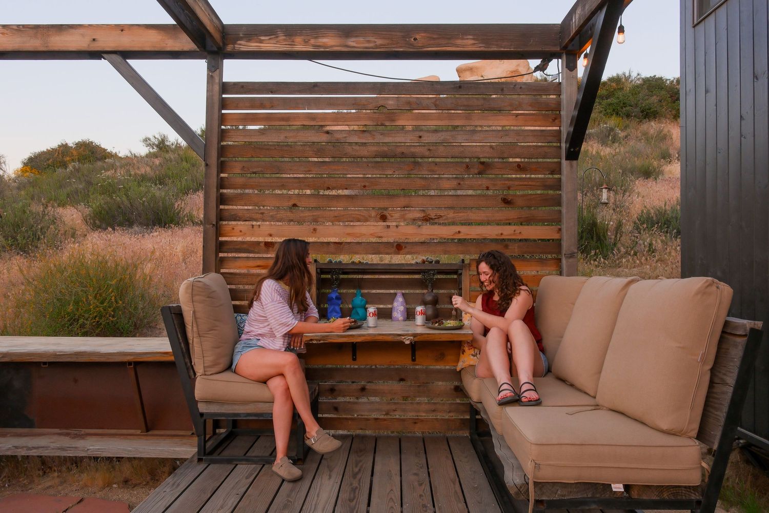 Two women sitting outdoors on a wooden deck patio with rustic fence, couch, and table at golden hour.