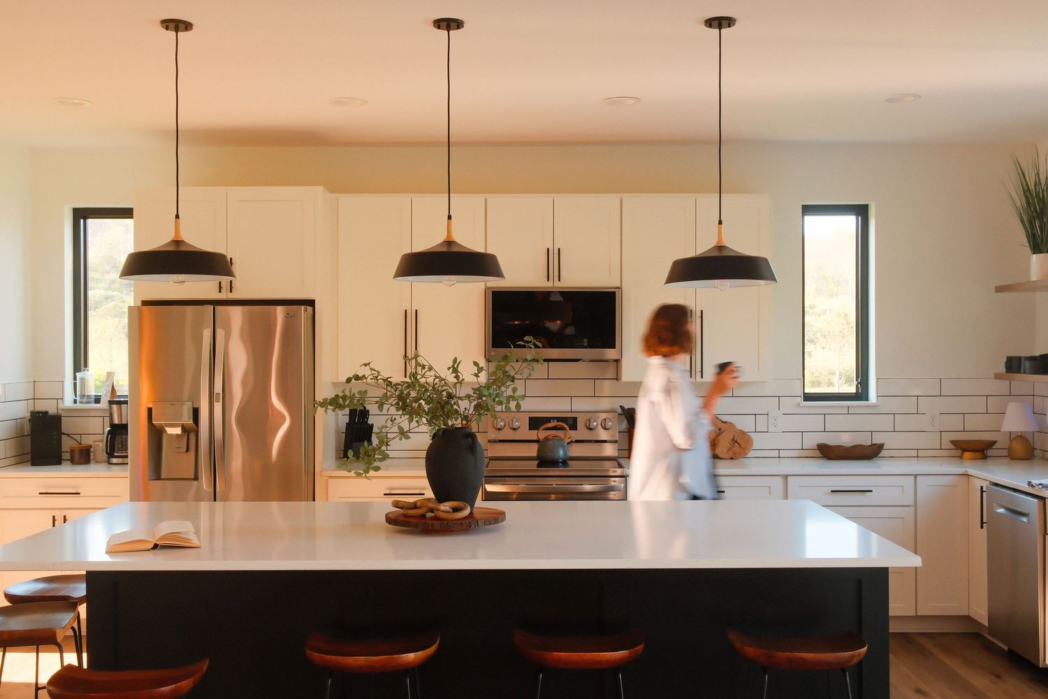 Modern white kitchen with black pendant lights, large island with bar stools, and stainless steel fridge.
