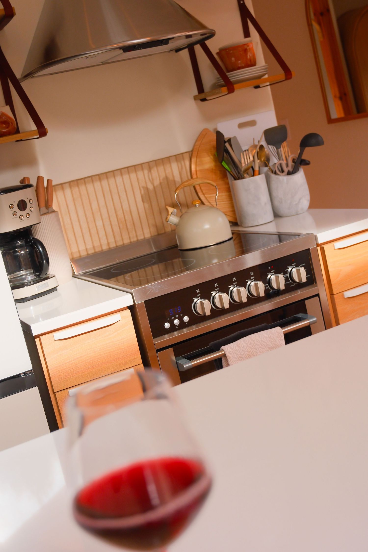 Cozy cabin kitchen with stainless range hood, induction stove, wooden cabinets and glass of red wine in foreground.